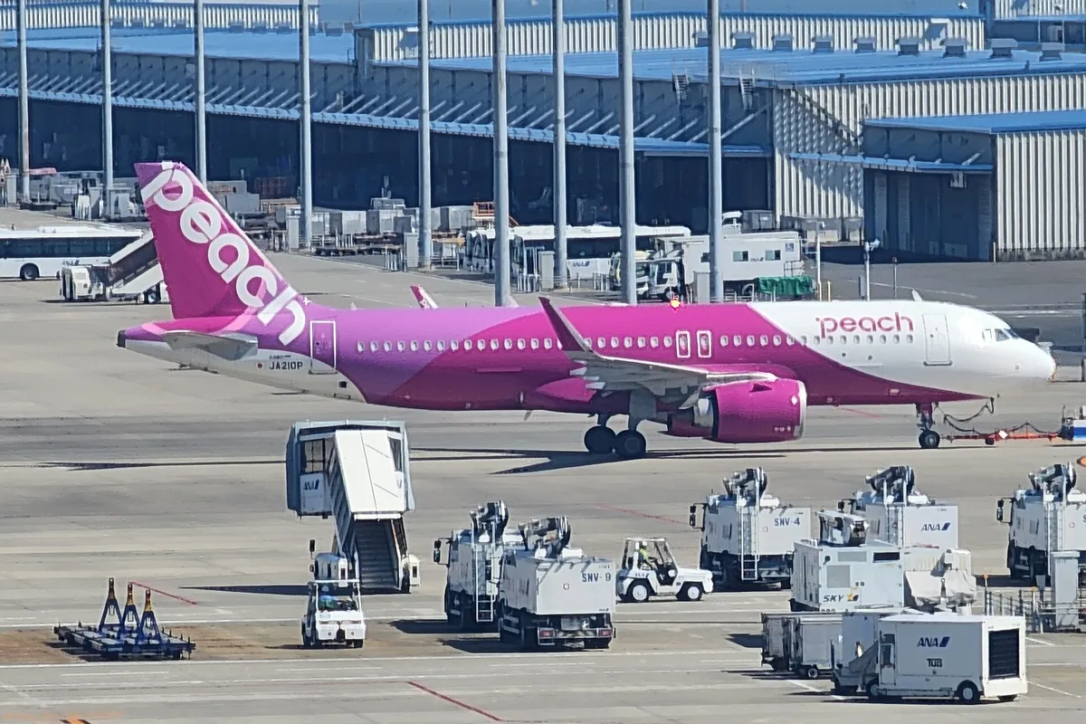 A pink Peach Aviation Airbus A320neo on the tarmac at a Japanese airport, surrounded by ground support vehicles and a jet bri