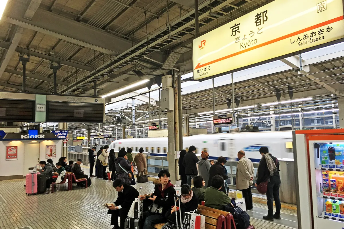 Passengers waiting on a Kyoto Station platform as a bullet train passes, with a Kyoto station sign and Kiosk shop overhead