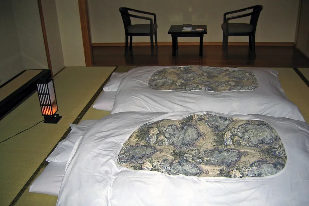 Two bedding sets with patterned covers and pillows laid out on a tatami mat floor, with chairs in the room