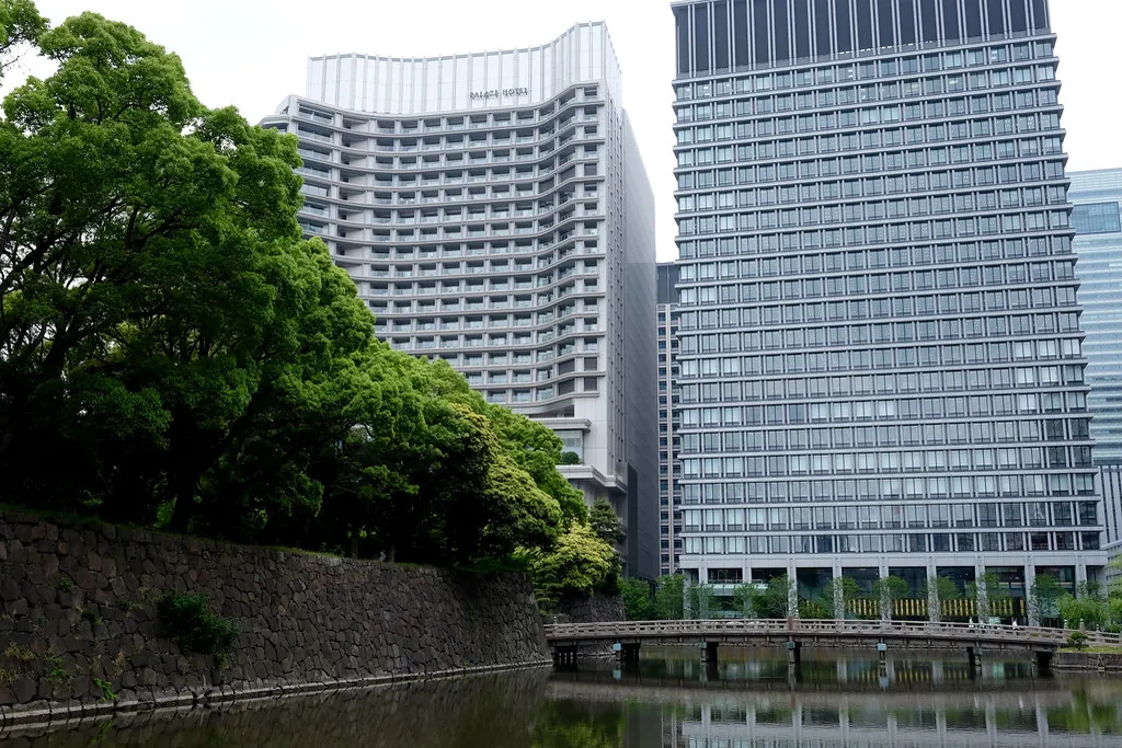 A modern glass-walled hotel building in Tokyo with trees, a stone wall, and a bridge over a moat