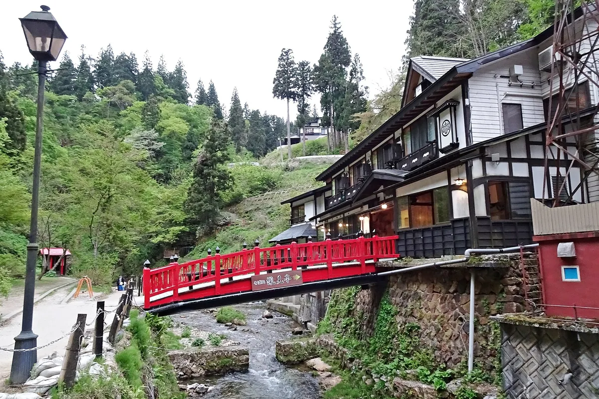 Wooden multi-story ryokan buildings along a river in Ginzan Onsen, with a red bridge and a street lamp