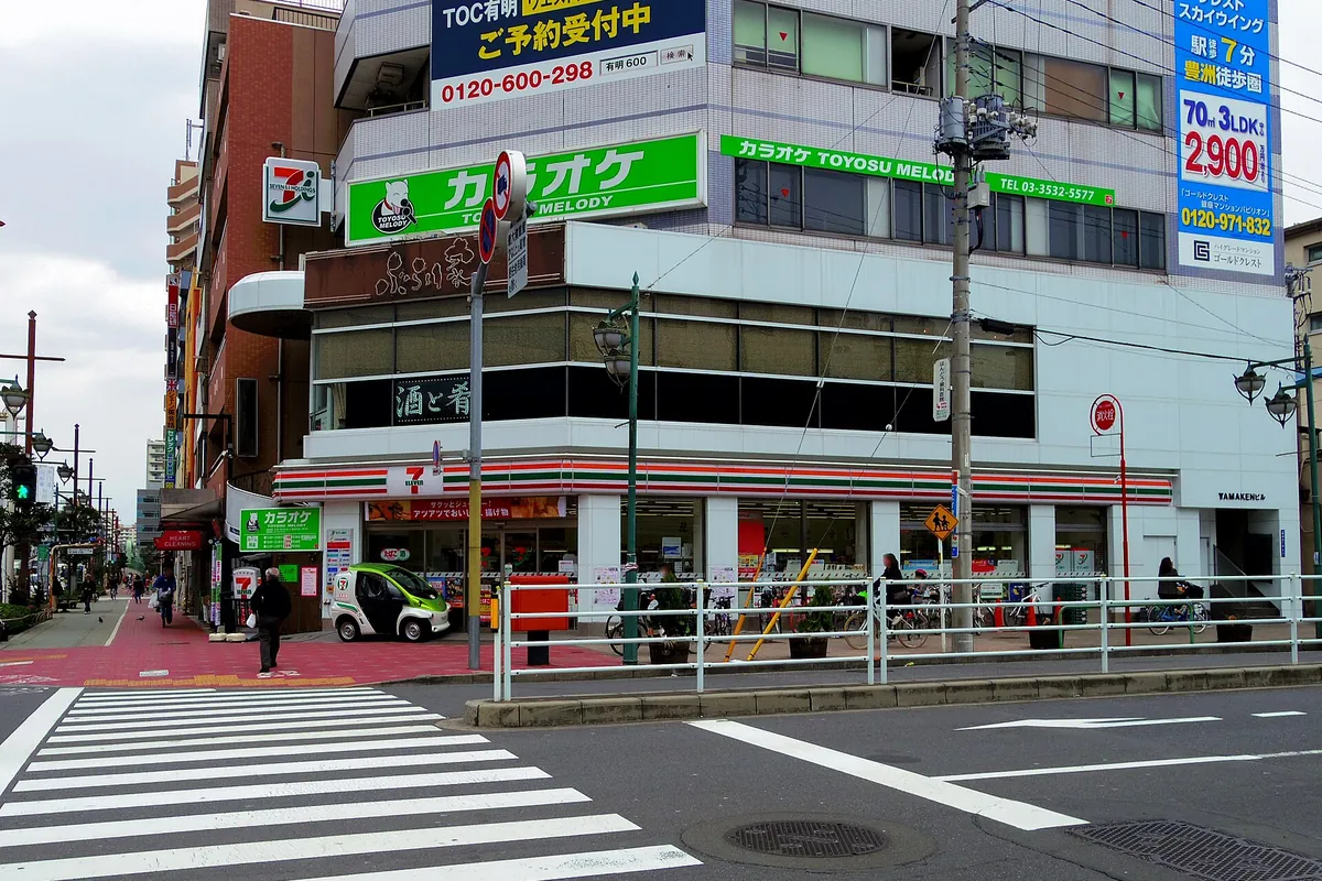 A 7-Eleven convenience store on a street corner in Tokyo, Japan, with a karaoke sign above and a crosswalk in front