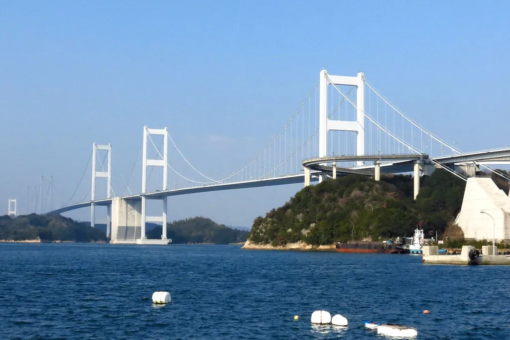 白い高い主塔を持つ来島海峡大橋が海にかかり、下には貨物船や係留された船が見える