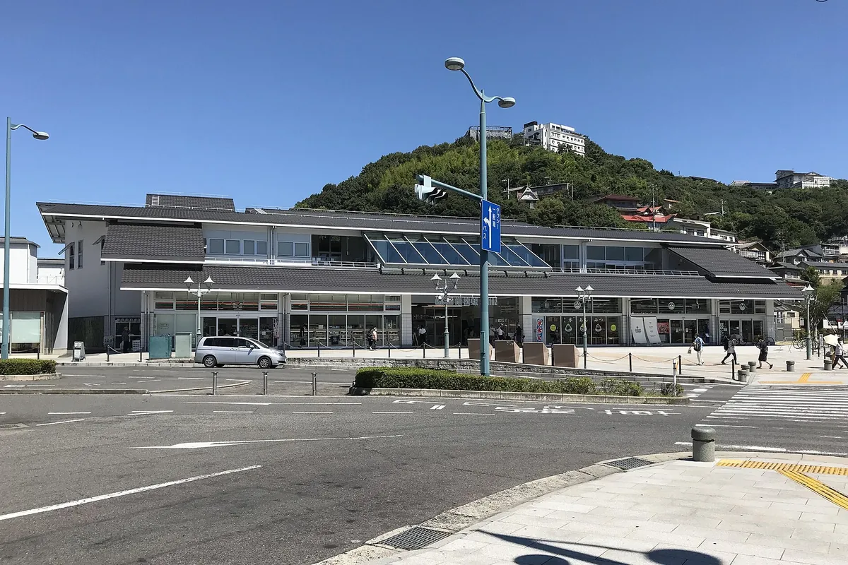 JR尾道駅の南口。瓦屋根の長い建物で、後ろには家が建つ木々に覆われた丘がある