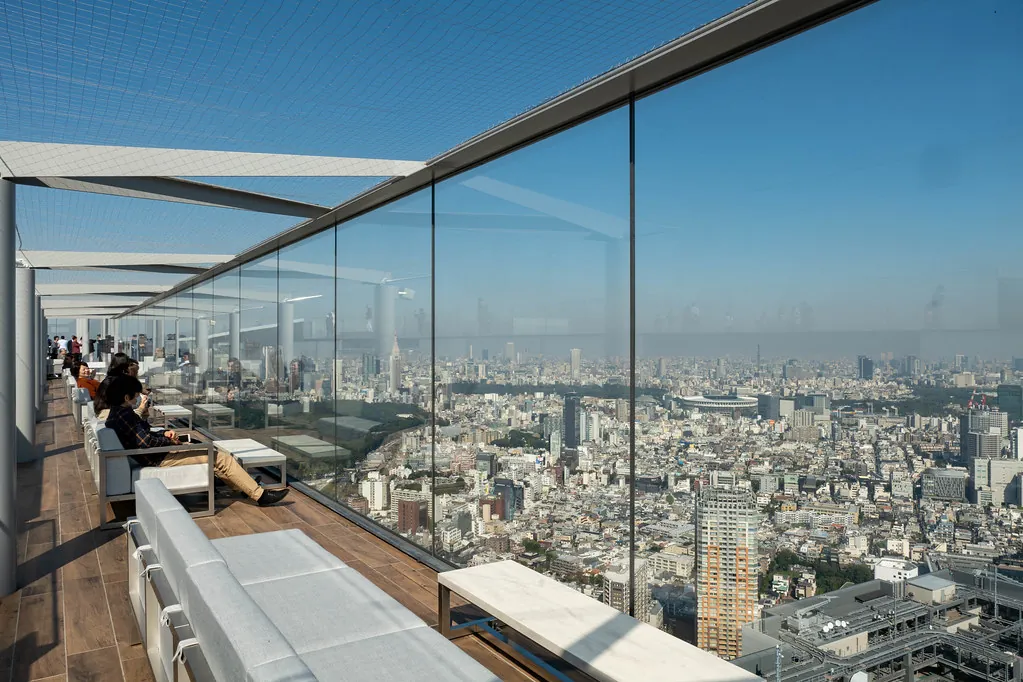 People sitting on benches on the open-air rooftop deck of SHIBUYA SKY with the Tokyo cityscape beyond the glass walls