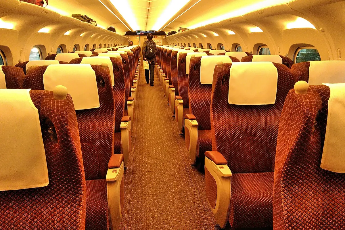 Interior of a Shinkansen Green Car with rows of red seats arranged two by two and a passenger walking down the aisle
