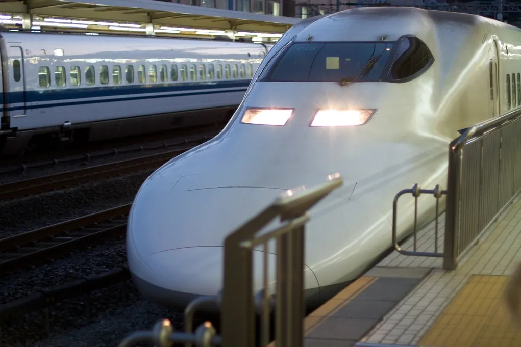 A white Shinkansen train with illuminated headlights arriving at a station platform, another train on the next track