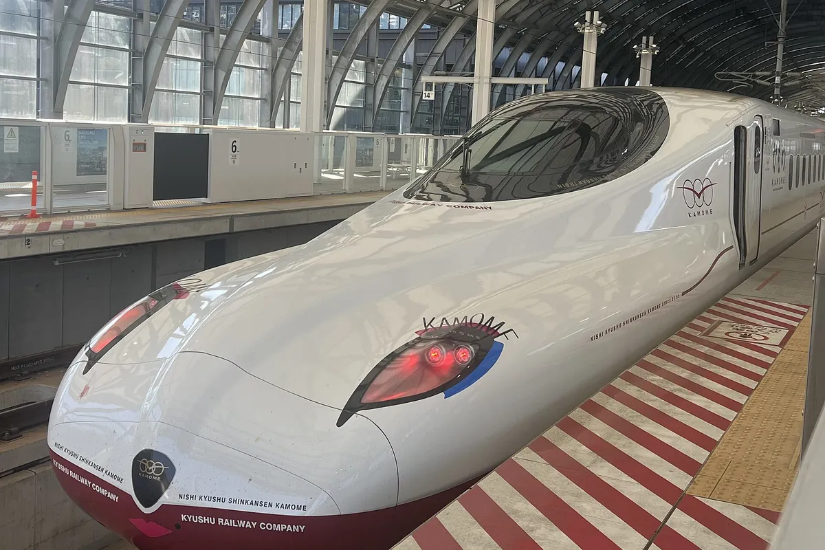 A white and red Kamome Shinkansen train at a station platform under an arched glass roof, with red and white striped platform