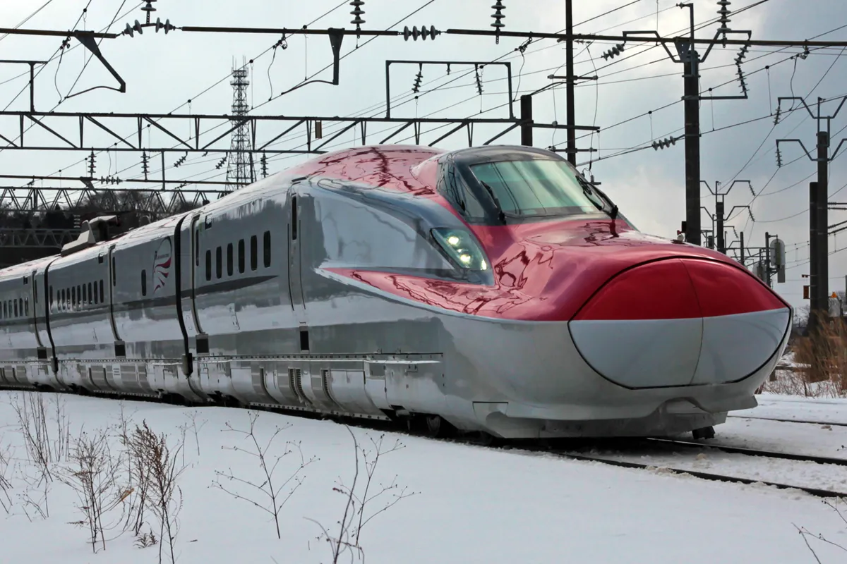 A red and silver E6 series Komachi Shinkansen train running through a snow-covered landscape