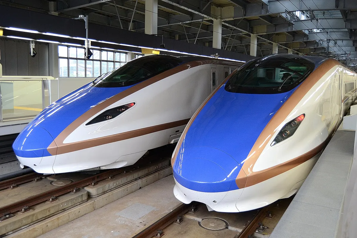 Two Hokuriku Shinkansen trains with blue, white, and gold stripes parked nose-to-nose at a station platform