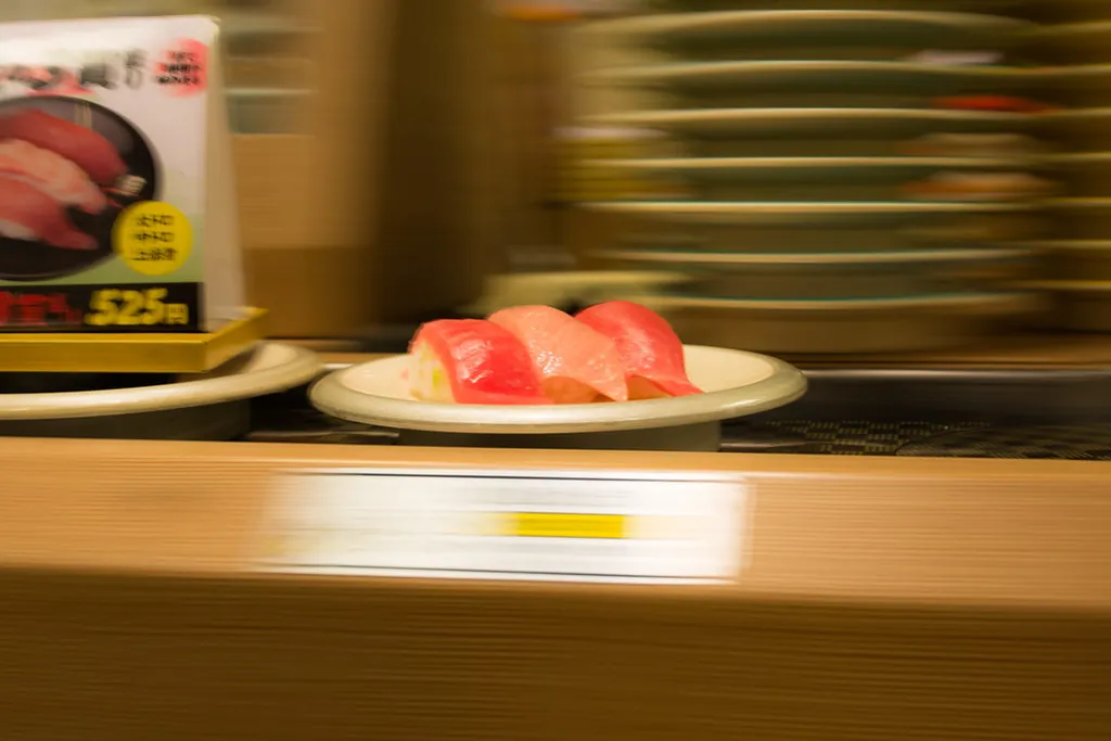 A plate of tuna sushi moving along a conveyor belt in a Japanese restaurant, with a menu sign showing a price in the backgrou