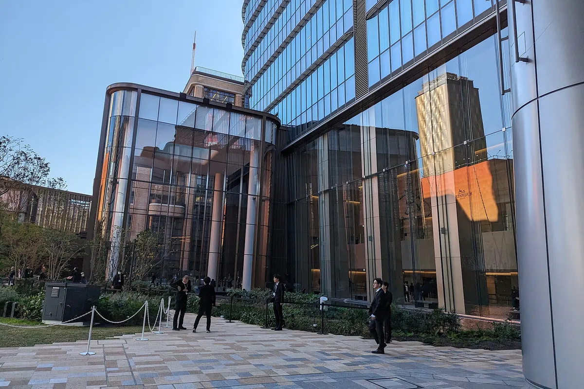 Curved glass facade of the Azabudai Hills complex in Minato, Tokyo, with people standing on the plaza outside
