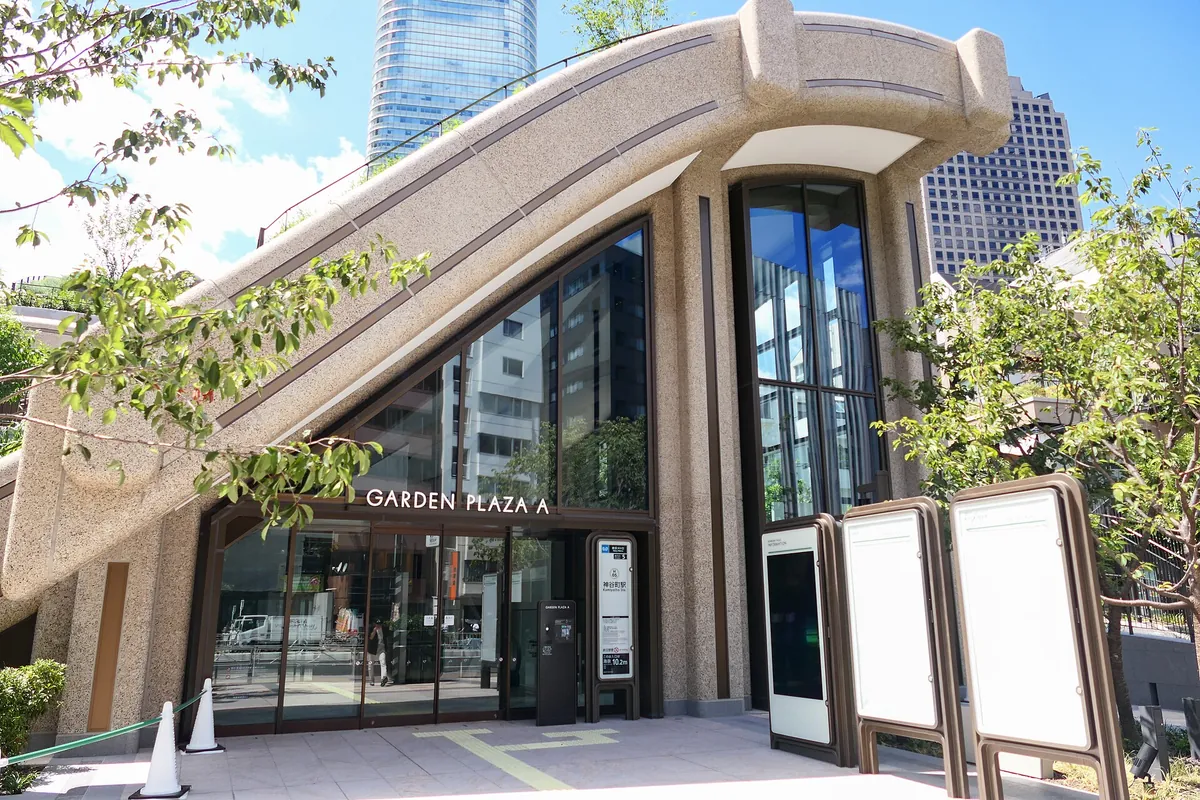 Entrance to Garden Plaza A at Azabudai Hills, a curved concrete building with a glass facade, trees, and a skyscraper behind