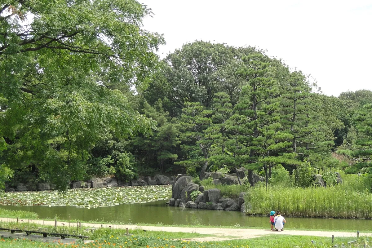 Pond with water lilies and large rocks at Nagai Botanical Garden in Osaka, with two children crouching on a path near trees