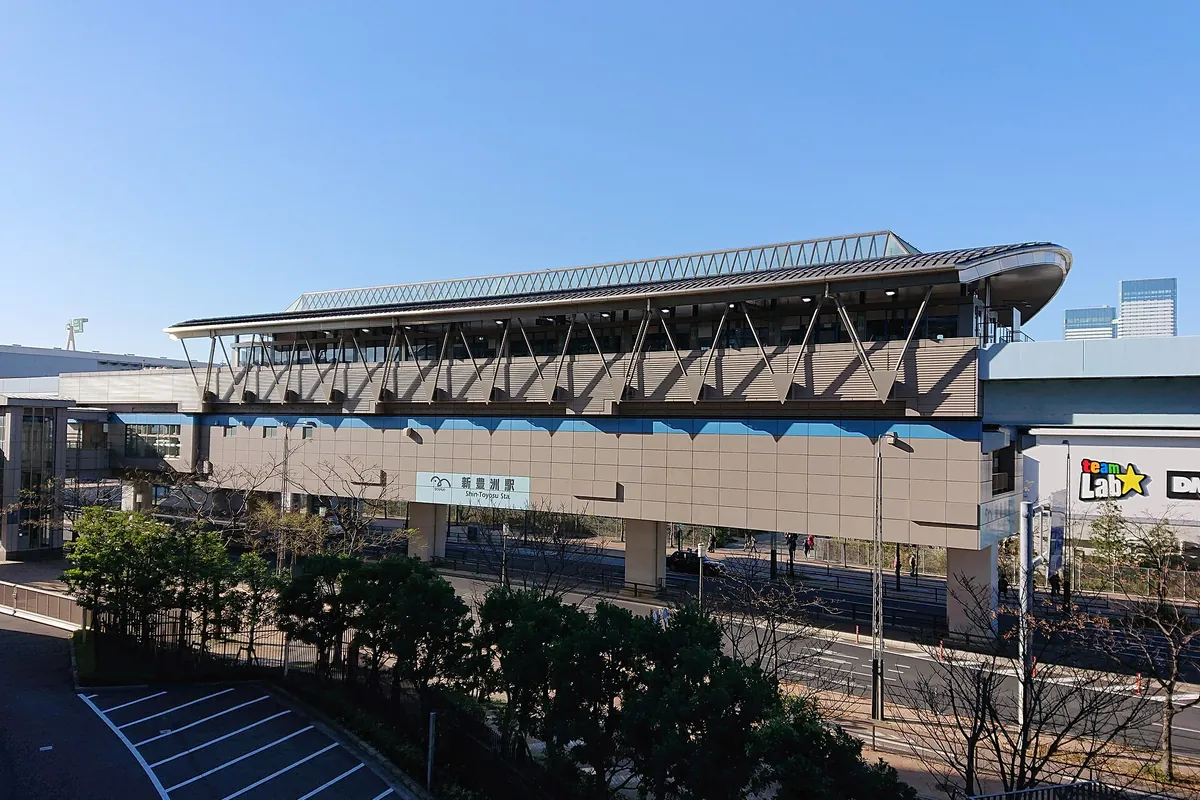 Elevated Shin-Toyosu Station building with its station sign, a parking lot in front, and a teamLab sign nearby