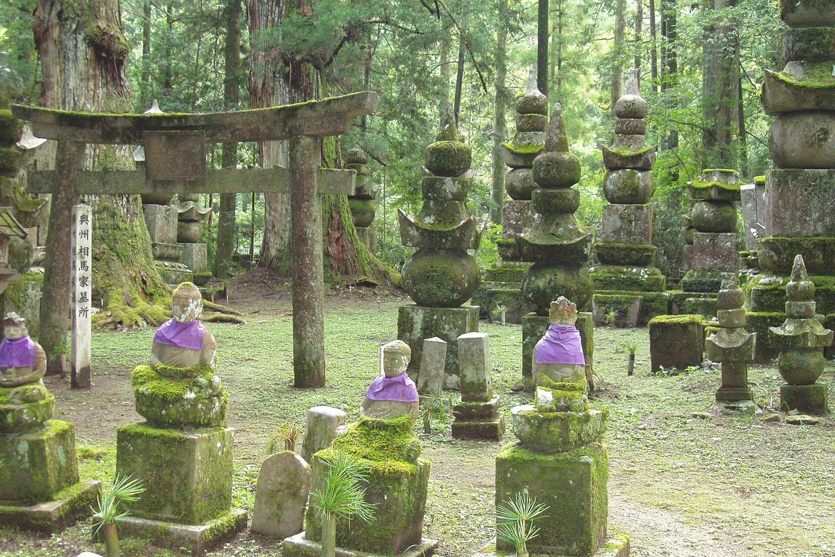 高野山・奥之院の杉並木の参道沿いに並ぶ苔むした石の鳥居、五輪塔、紫の前掛けをした地蔵