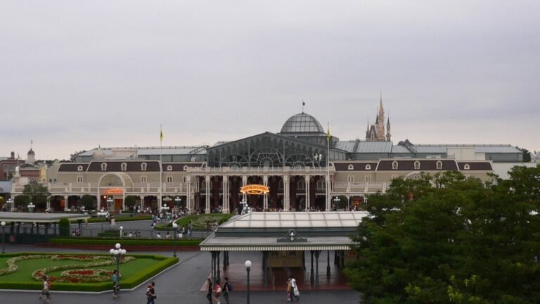 The Tokyo Disneyland Main Entrance gate building seen from above, with a plaza, flower bed, and guests walking toward it