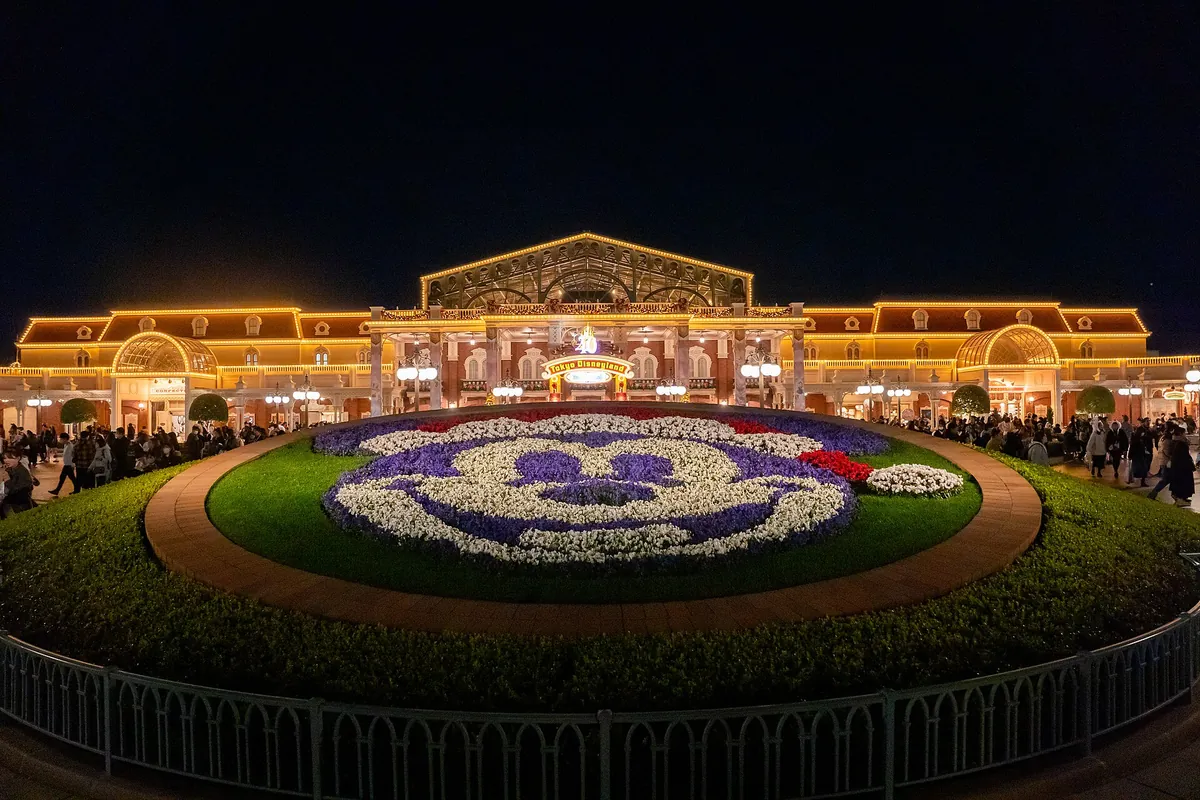 Tokyo Disneyland Main Entrance at night with a Mickey-shaped flower bed in front of the illuminated station-style building