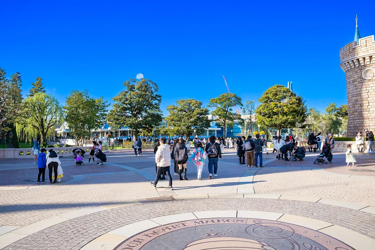 A crowd gathered in the plaza near Tokyo Disneyland's castle, with a stone turret on the right and a round plaque on the pave