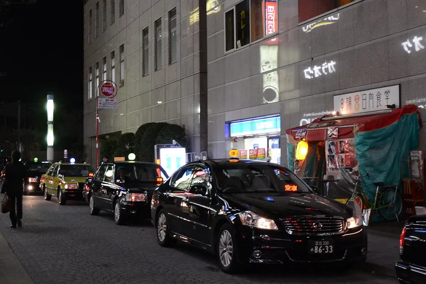 東京夜晚的街道，計程車沿著路邊排隊，店家招牌亮著燈，旁邊有一座有頂棚的小吃攤