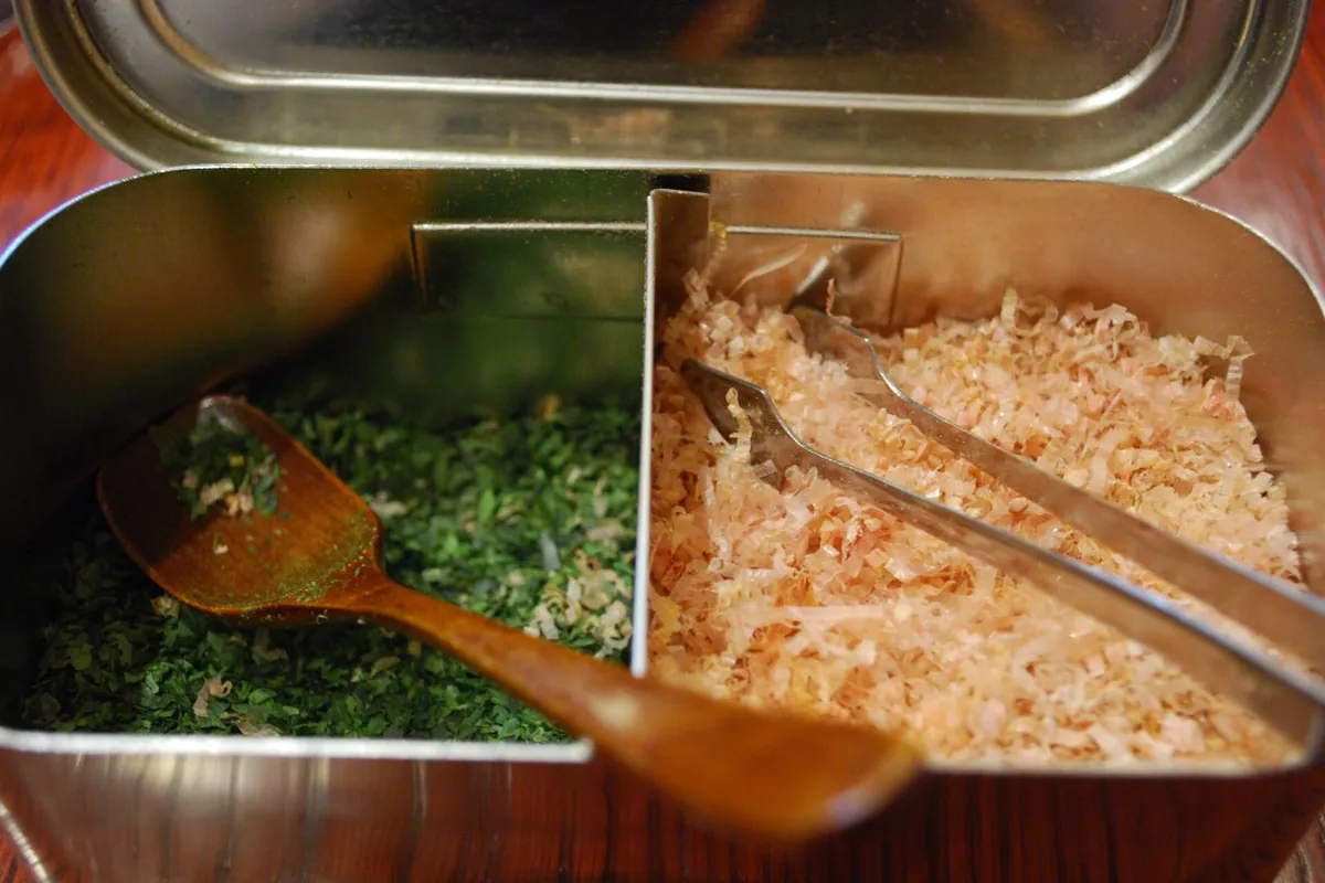 A tray of shaved dried bonito flakes (katsuobushi) next to chopped nori seaweed with serving tongs