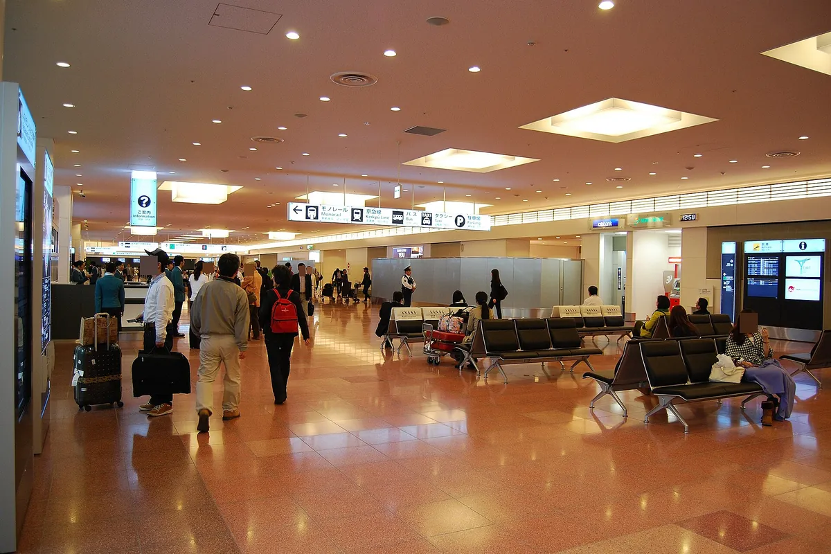 Travelers with suitcases walking through a wide Haneda Airport International Terminal hall past rows of black waiting chairs