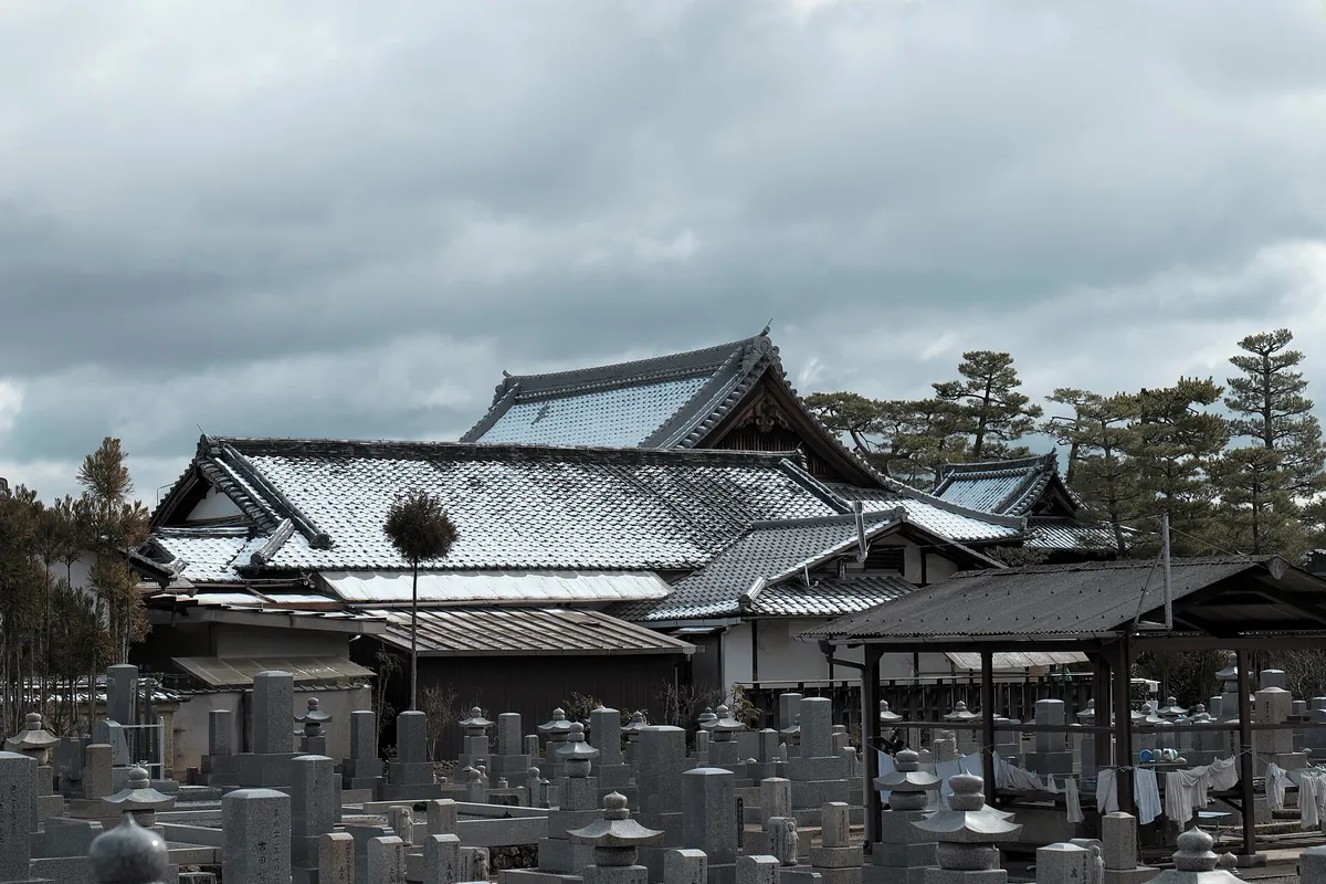 Snow-dusted tiled roofs of a temple in Kyoto with rows of stone gravestones in the foreground