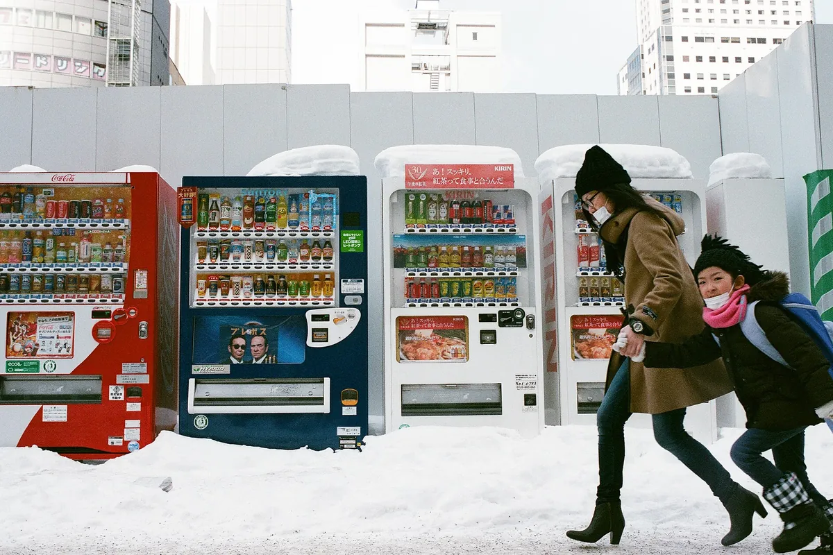 A woman and child wearing masks walking past vending machines on a snow-covered street in Sapporo, Japan